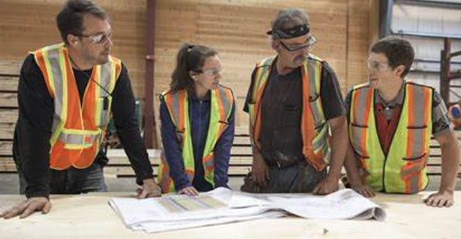 Mark Eyjolfson stands at a table with plans on it, talking to 3 coworkers. Everyone is wearing high-visibility vests and safety glasses.