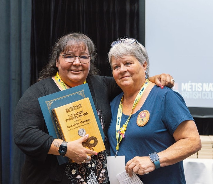 Two people stand smiling at the camera. One holds an award plaque.