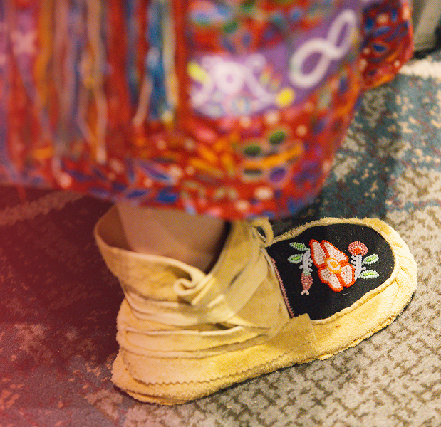 A yellow-coloured moccasin with a red- and green-coloured beaded flower on a black background and the hem of a colourful skirt.