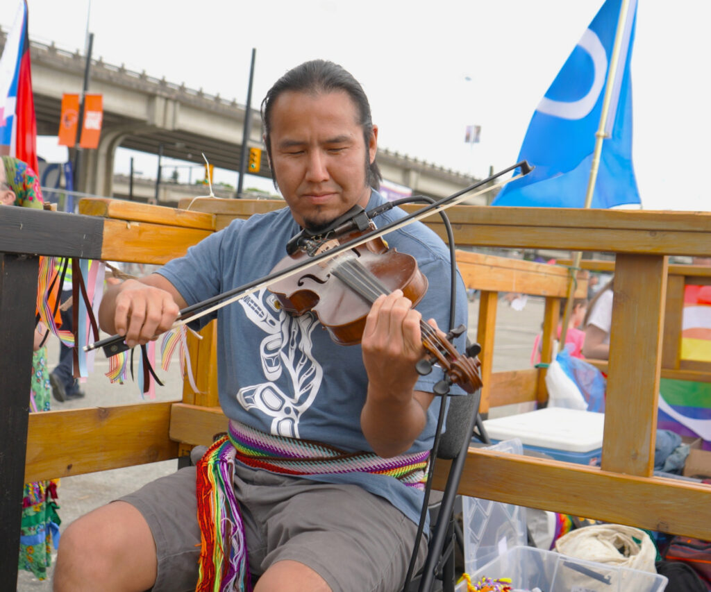 A man playing the fiddle is sitting on a wooden structure on the Pride parade float.