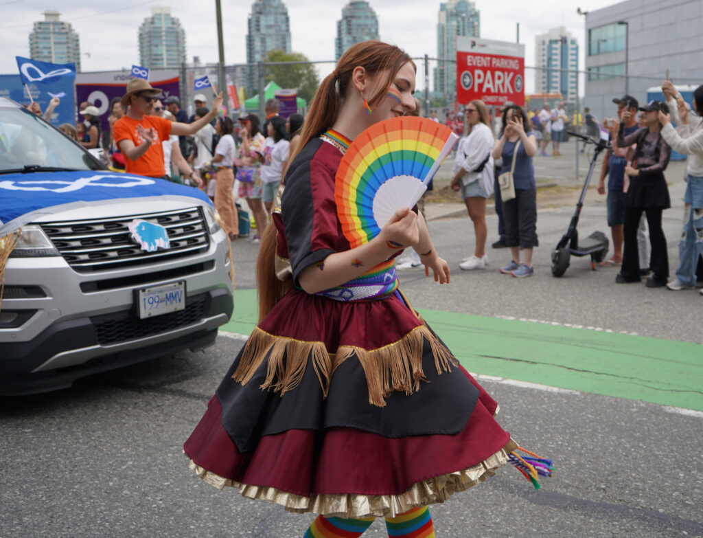A woman wearing a traditional Métis jigging outfit holding a rainbow fan while walking in the Pride parade.