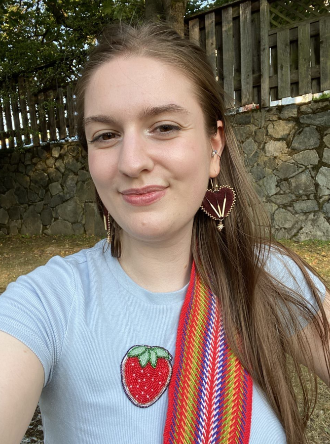 Seone Anderson is smiling in a grey shirt with a red Métis sash and a beaded strawberry pin against an outdoor background.