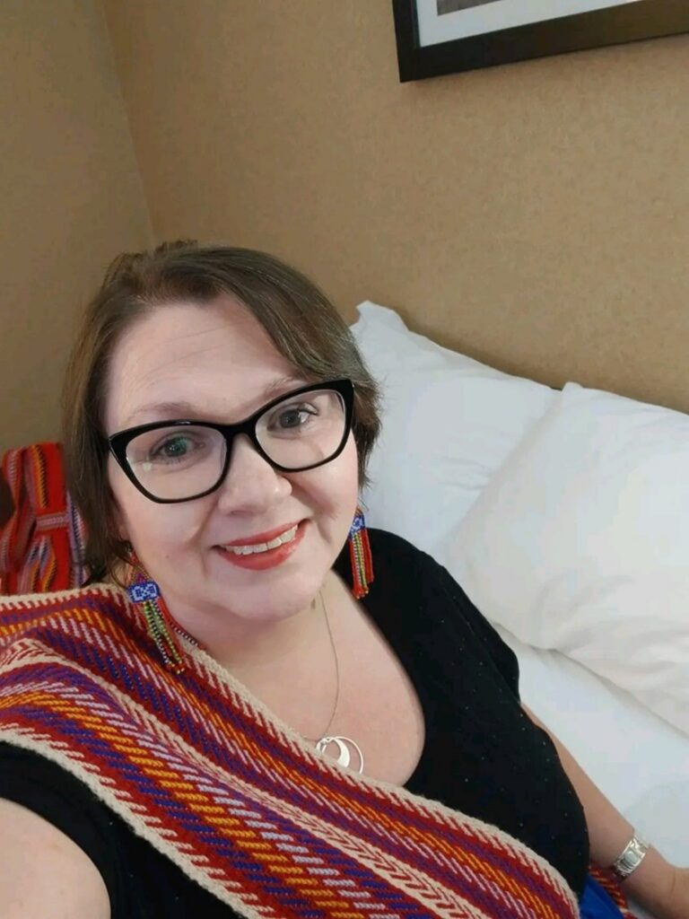 Melissa Kelm is smiling in a black shirt with a red Métis sash draped over her shoulder against an indoor background.