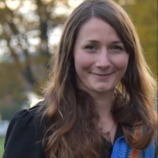 Mia Yule is smiling in a black shirt with a blue Métis sash draped over her shoulder against an outdoor background.