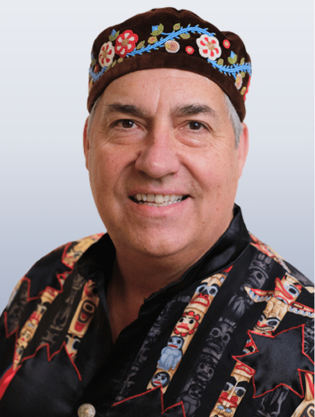 Louis De Jaeger is smiling in a black shirt with embroidered totem poles and is also wearing a black hat with beaded flowers on a white background.