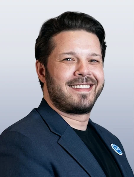 Anthony Wingham is smiling in a navy blue blazer with a Métis pin on his lapel on a grey background.