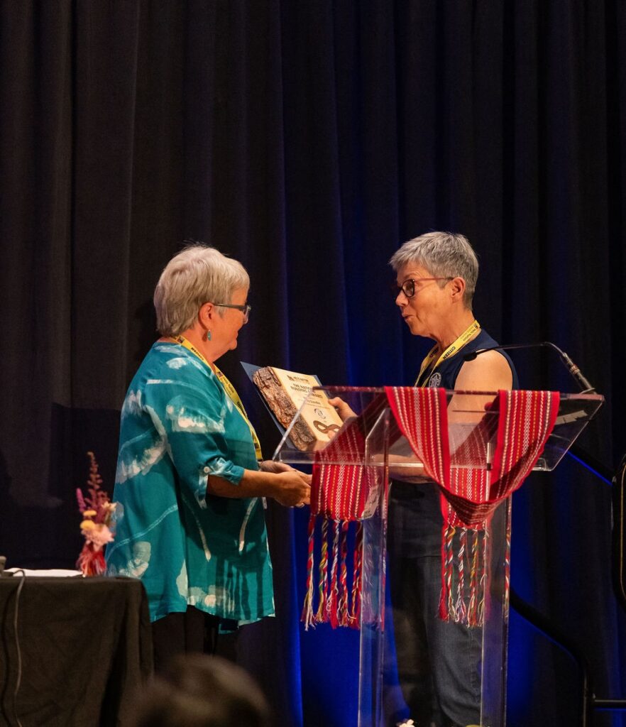 Two middle aged women stand at a podium. One is receiving a wooden award plaque.