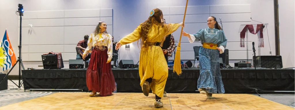 A group of women wearing traditional Métis clothing, one passing a broom to the other on a dance floor.