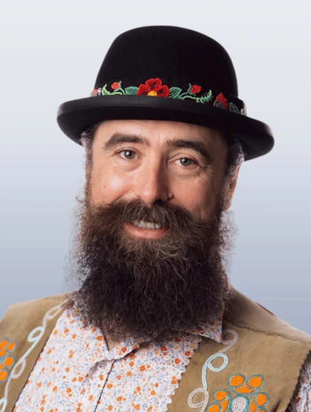 Patrick Harriott is smiling in a paisley shirt with a beaded vest and a black beaded hat against a white background.