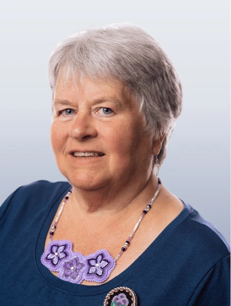 Susie Hooper is smiling in a blue shirt, wearing a purple beaded necklace and a beaded pin against a white background.